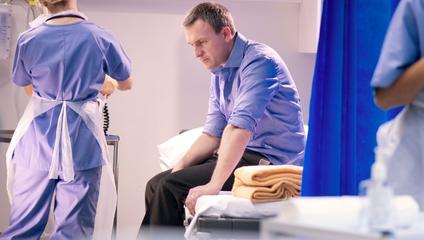 Image of patient sitting on the side of hospital bed with doctor in the background to close the gap on psychiatric emergencies